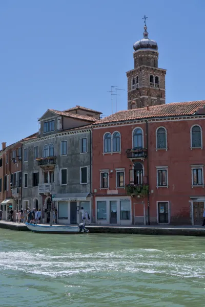 Architektonische Ansicht eines Gebäudes auf Murano am Kanal, mit einem Kirchturm im Hintergrund, aufgenommen im Hochformat.