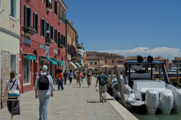 Ljudi šetaju duž sunčanog šetališta na ostrvu Murano, Italija, sa šarenim kućama i brodovima pored vode.
