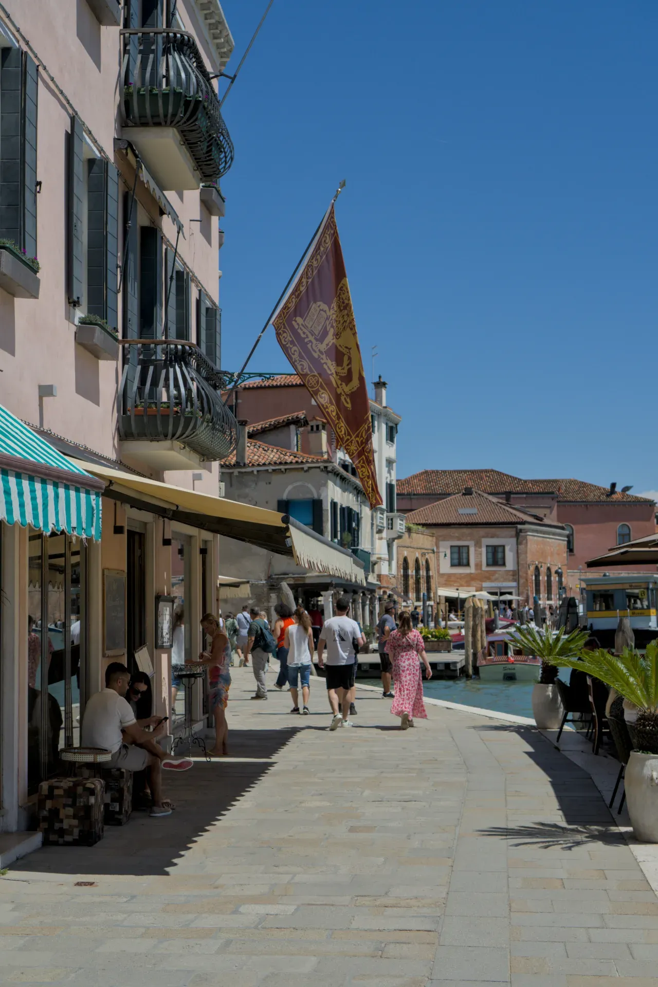 People stroll along a canal on the island of Murano, Venice, under a waving Venetian flag.