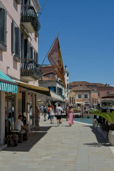 Menschen schlendern entlang eines Kanals auf der Insel Murano, Venedig, unter einer wehenden venezianischen Flagge.