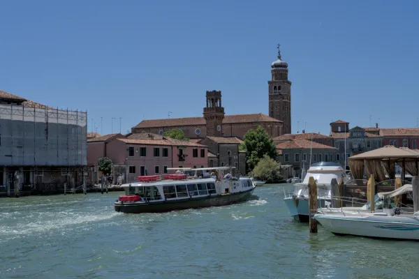 Saobraćaj brodova na kanalu ostrva Murano, Venecija, sa istorijskim crkvenim zgradama u pozadini na suncu.