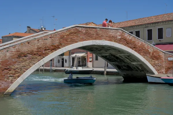 Kameni most sa opečnim akcentima premošćuje kanal na ostrvu Murano, Italija, sa čamcima ispod mosta i parom na parapetu.