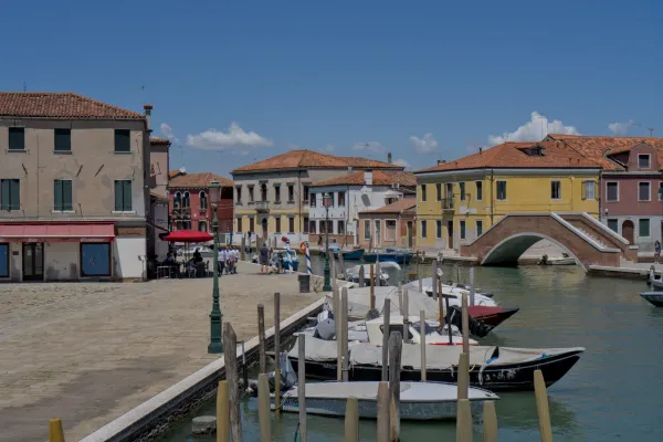 Ostrvo Murano u Veneciji prikazuje kanal sa čamcima i tradicionalni kameni most, okružen šarenim kućama.