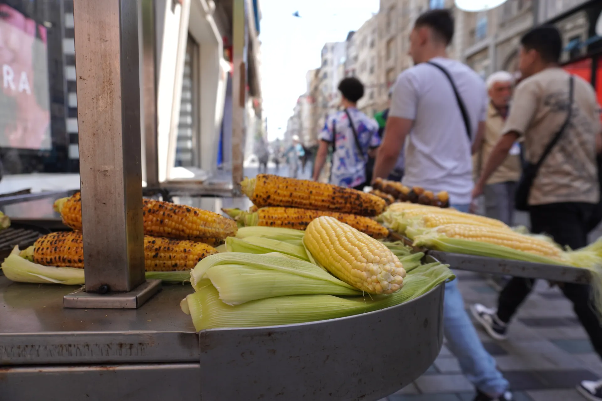 A street food cart in Istanbul filled with grilled corn on the cob and fresh corn cobs, with people walking by in the background.