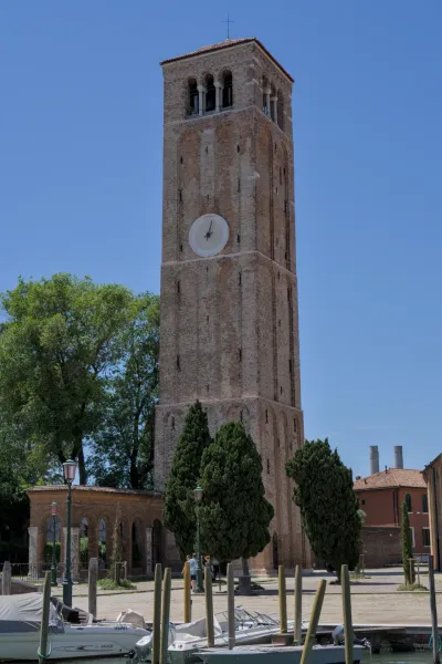 Cigleni zvonik ostrva Murano pod plavim nebom, sa drvećem i vodom ispred.