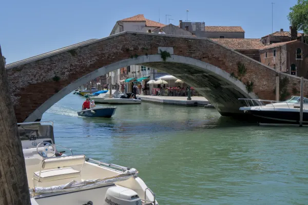 Istorijski kameni most na ostrvu Murano, Venecija, koji premošćuje kanal sa malim čamcima i kafićima.