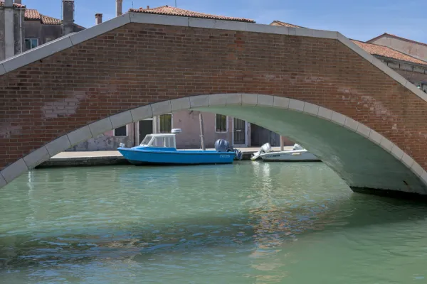 Crveni kameni most od cigle sa kamenim ukrasima premošćuje kanal, na kome leži plavi čamac, u blizini ostrva Murano.