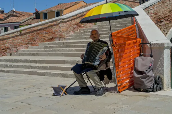 Ein älterer Mann spielt Akkordeon auf der Insel Murano, Venedig, Italien, unter einem bunten Regenschirm.