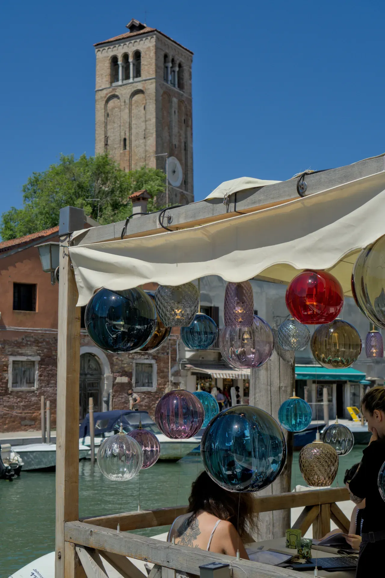 Handcrafted Murano glass lights hang over a canal in Venice with a tower in the background.