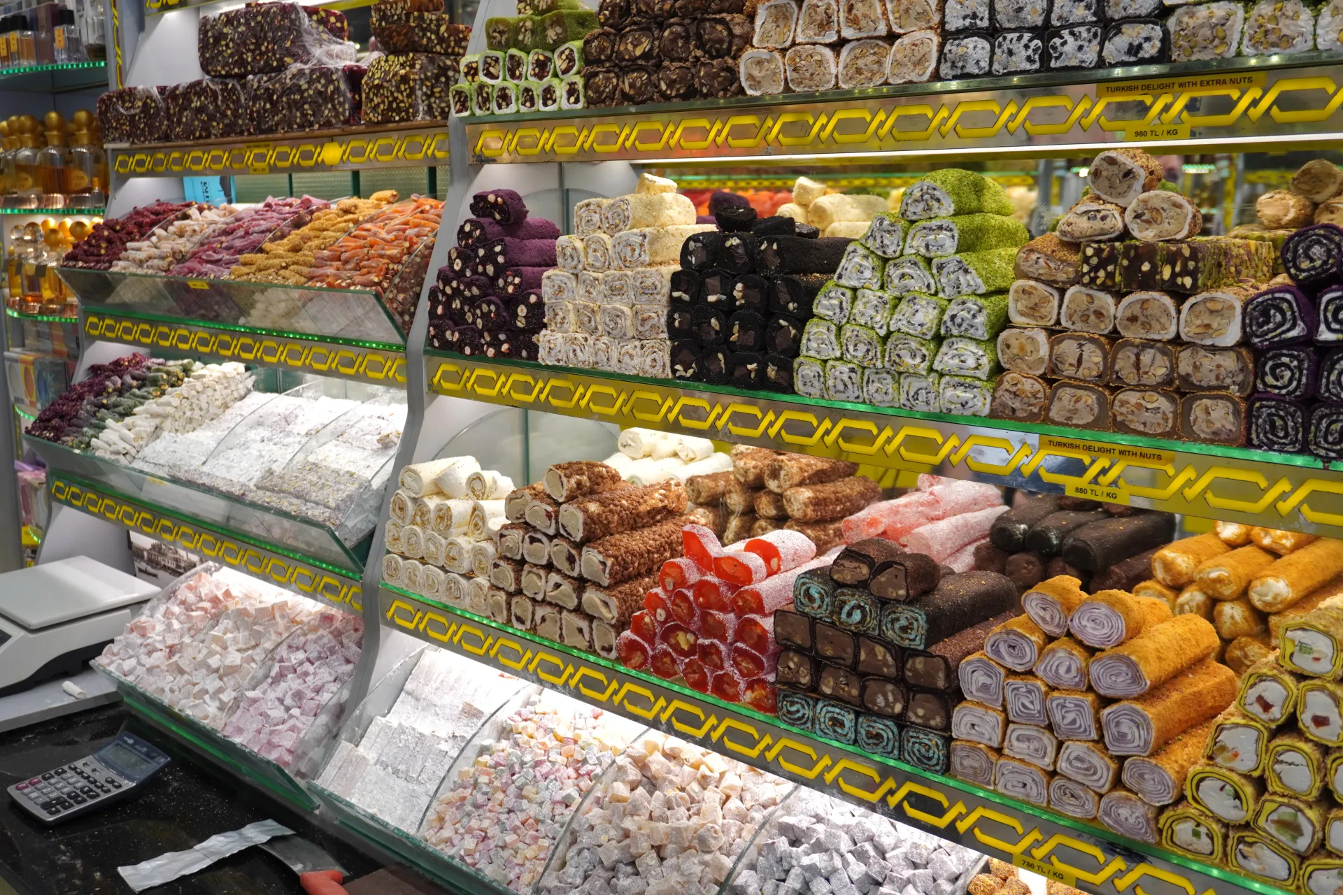 A well-lit shop interior in Istanbul displays rows of various types of Turkish Delight, including cubed and rolled varieties, some dusted with powdered sugar and others filled with nuts, presented in glass cases.