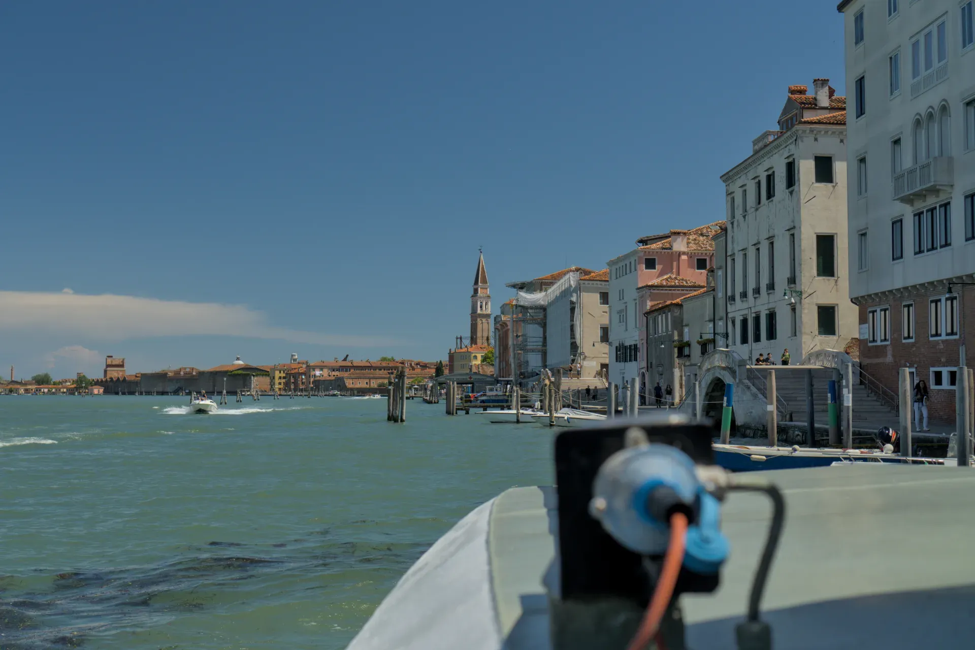 A sunny day in Venice, view of the canal bank with traditional buildings reflected in the turquoise water, and a passing boat.