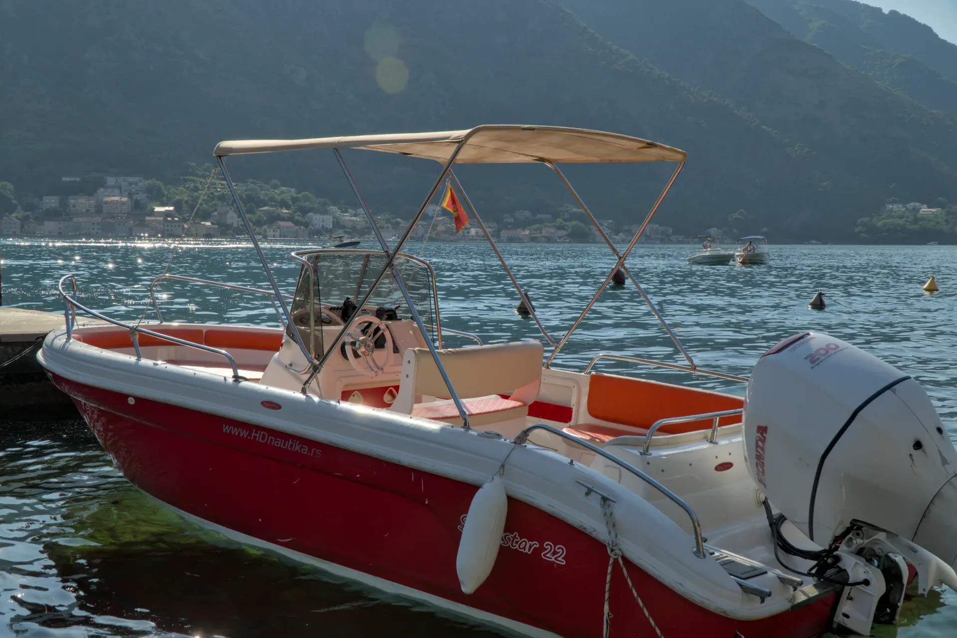 A red and white motorboat rests in the sparkling water of a harbor in Kotor, Montenegro, with green hills in the background.
