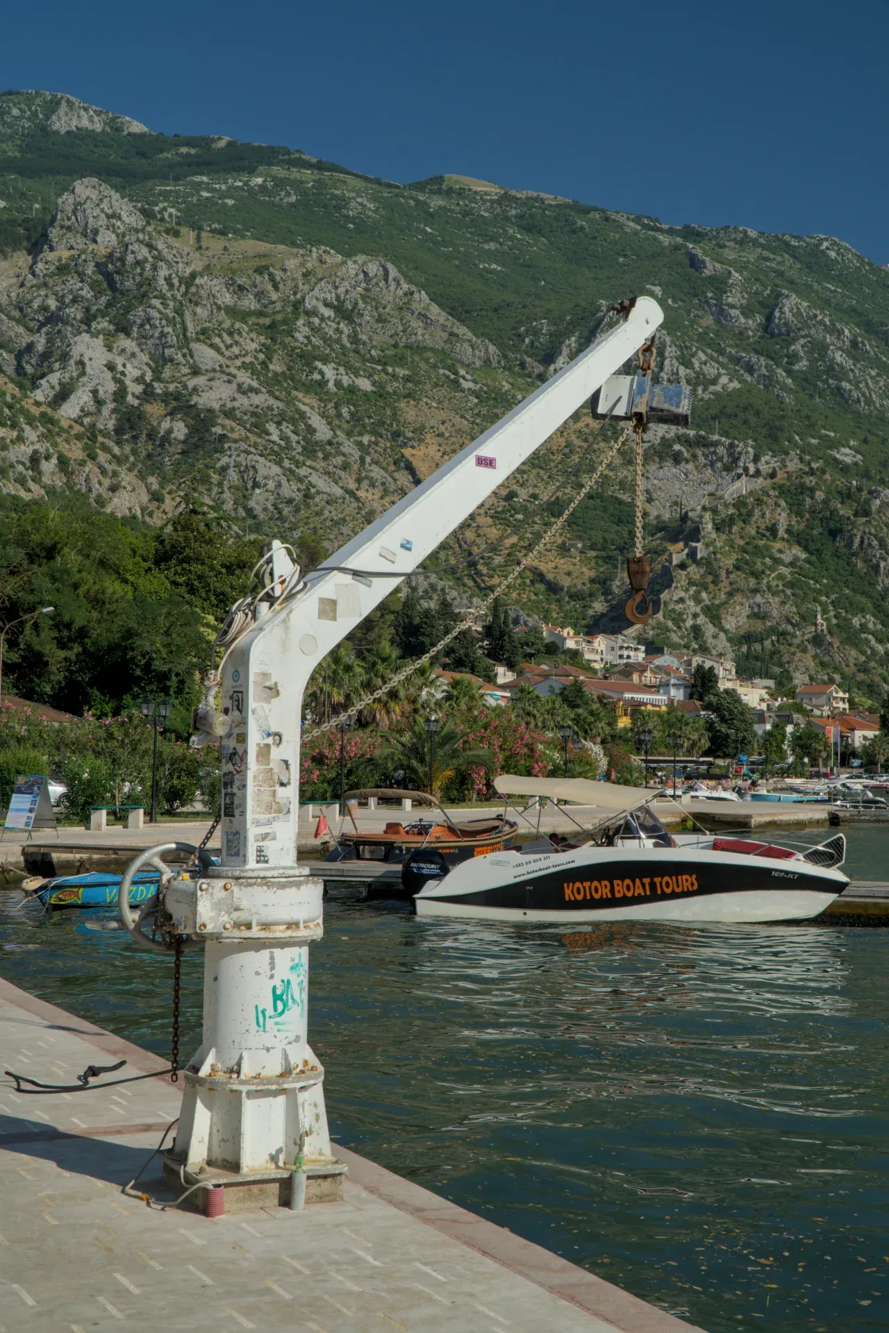 A white harbor crane stands in the foreground, with boats and the city of Kotor in the background, against a mountainous landscape.