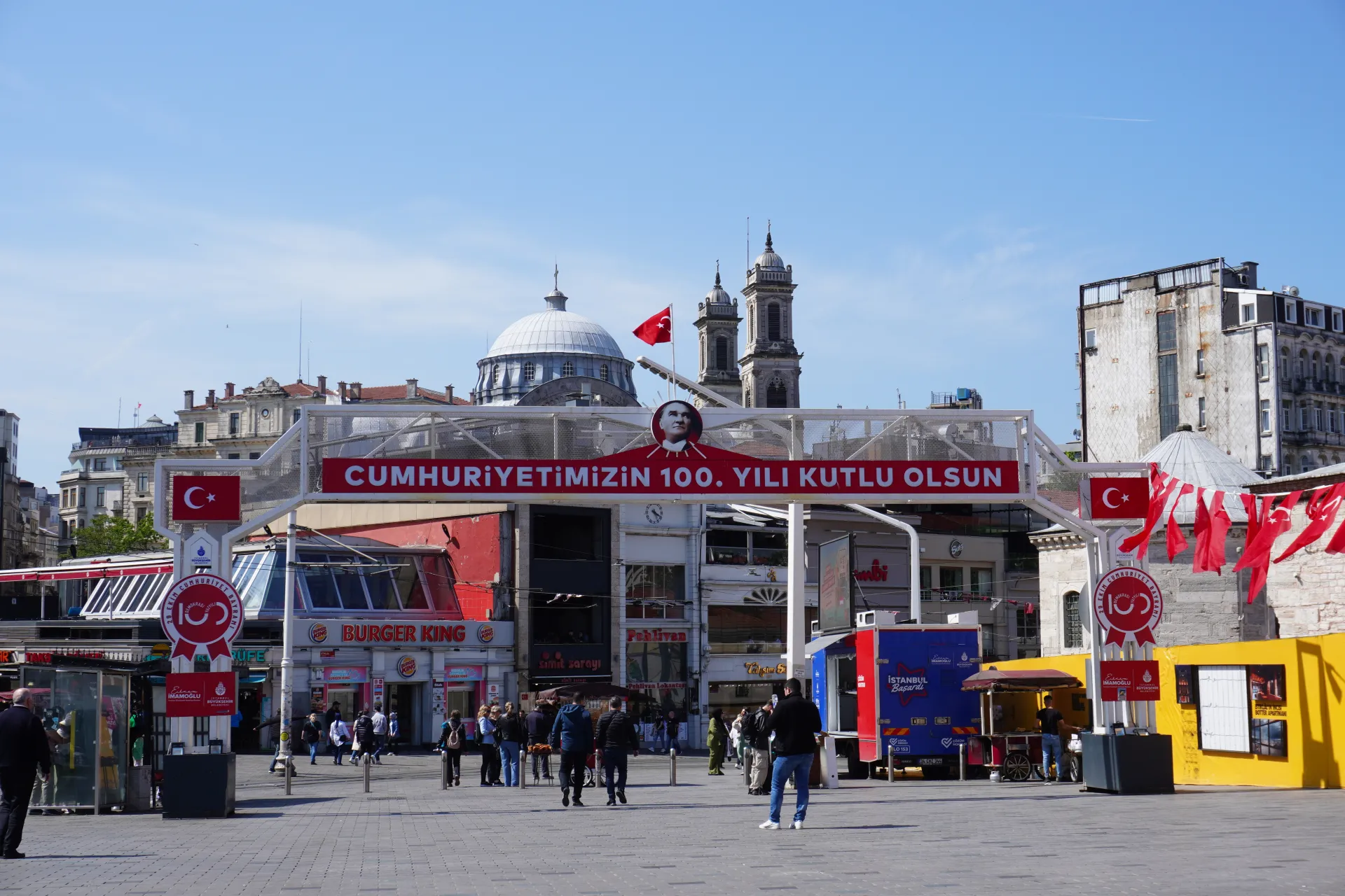 A view of Taksim Square in Istanbul, Turkey, adorned with a banner celebrating the 100th anniversary of the Republic, featuring a portrait of Atatürk and numerous Turkish flags, with people and buildings in the background under a clear blue sky.