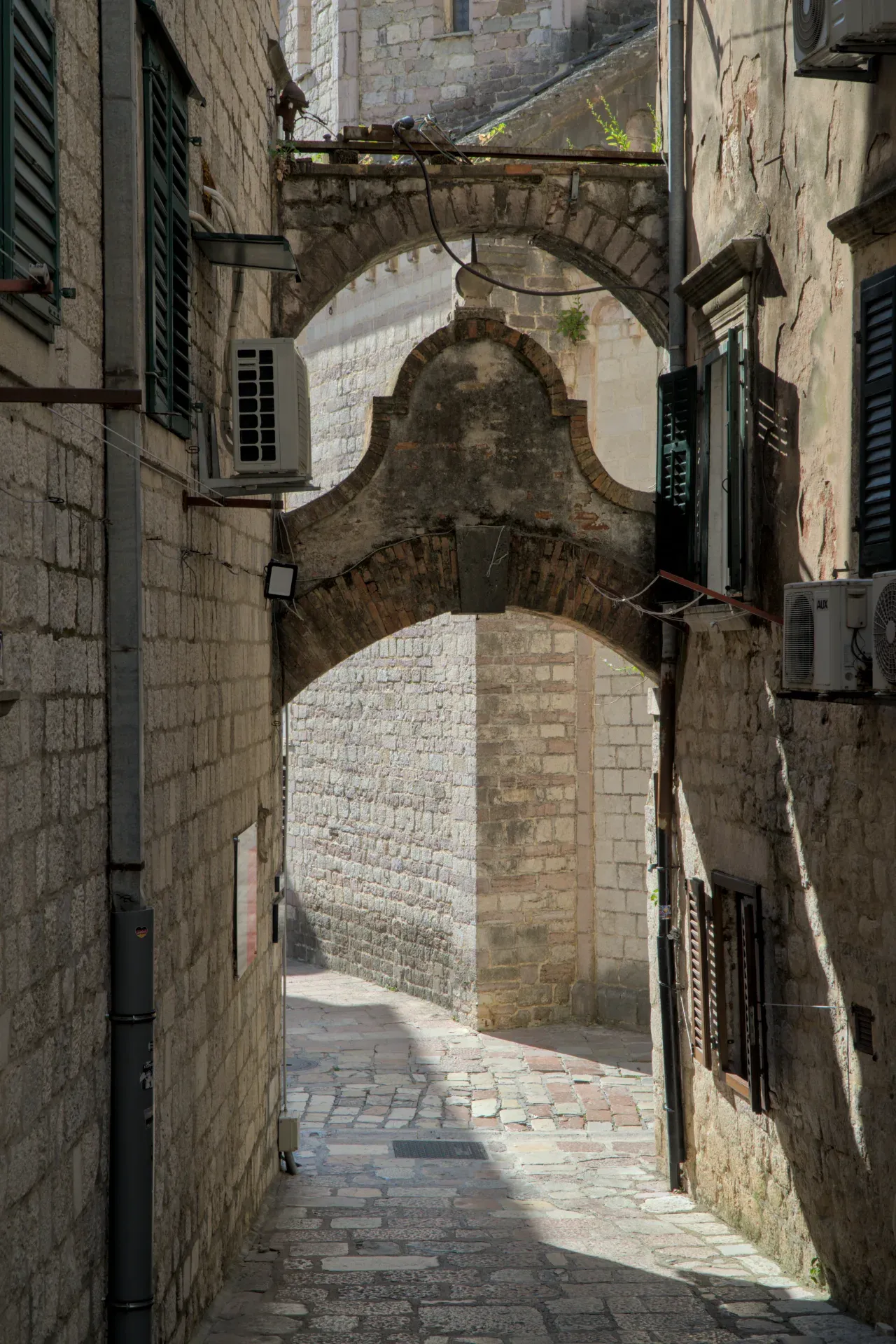 Old stone archways in a narrow alley in Kotor, Montenegro, with sunshine and shadows.