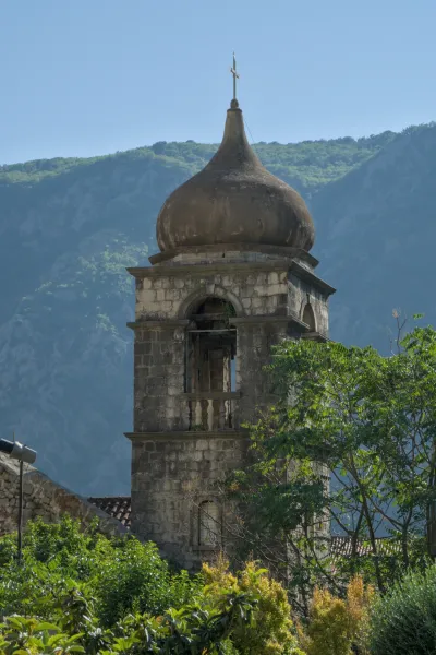 Clocher historique en pierre avec dôme et croix, sur fond de montagnes boisées et de ciel bleu à Kotor.