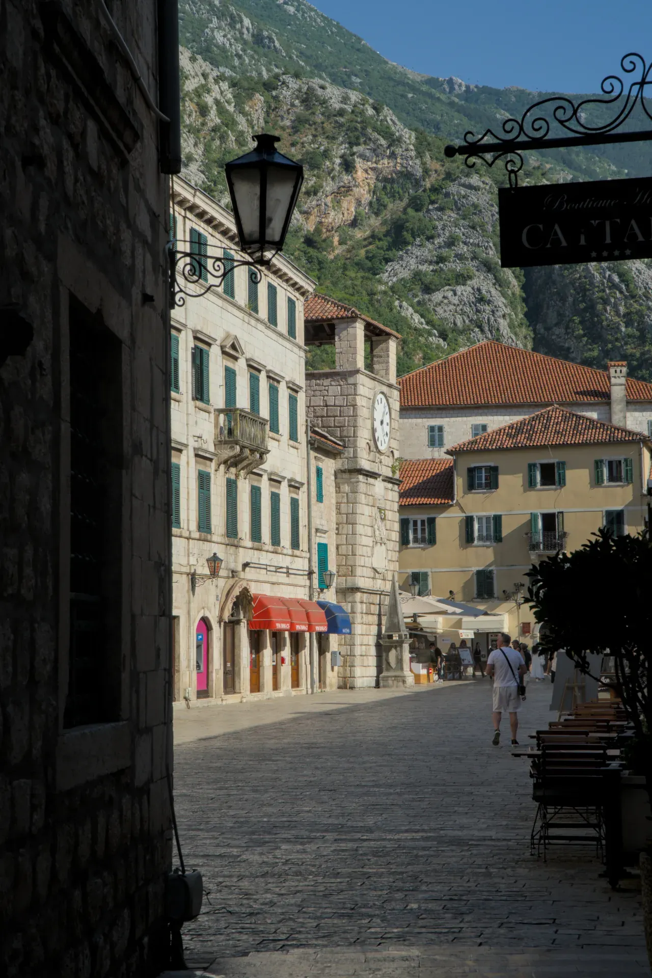 Toma pictórica de un callejón en Kotor con una torre del reloj histórica, enmarcada por edificios antiguos y paisaje montañoso.