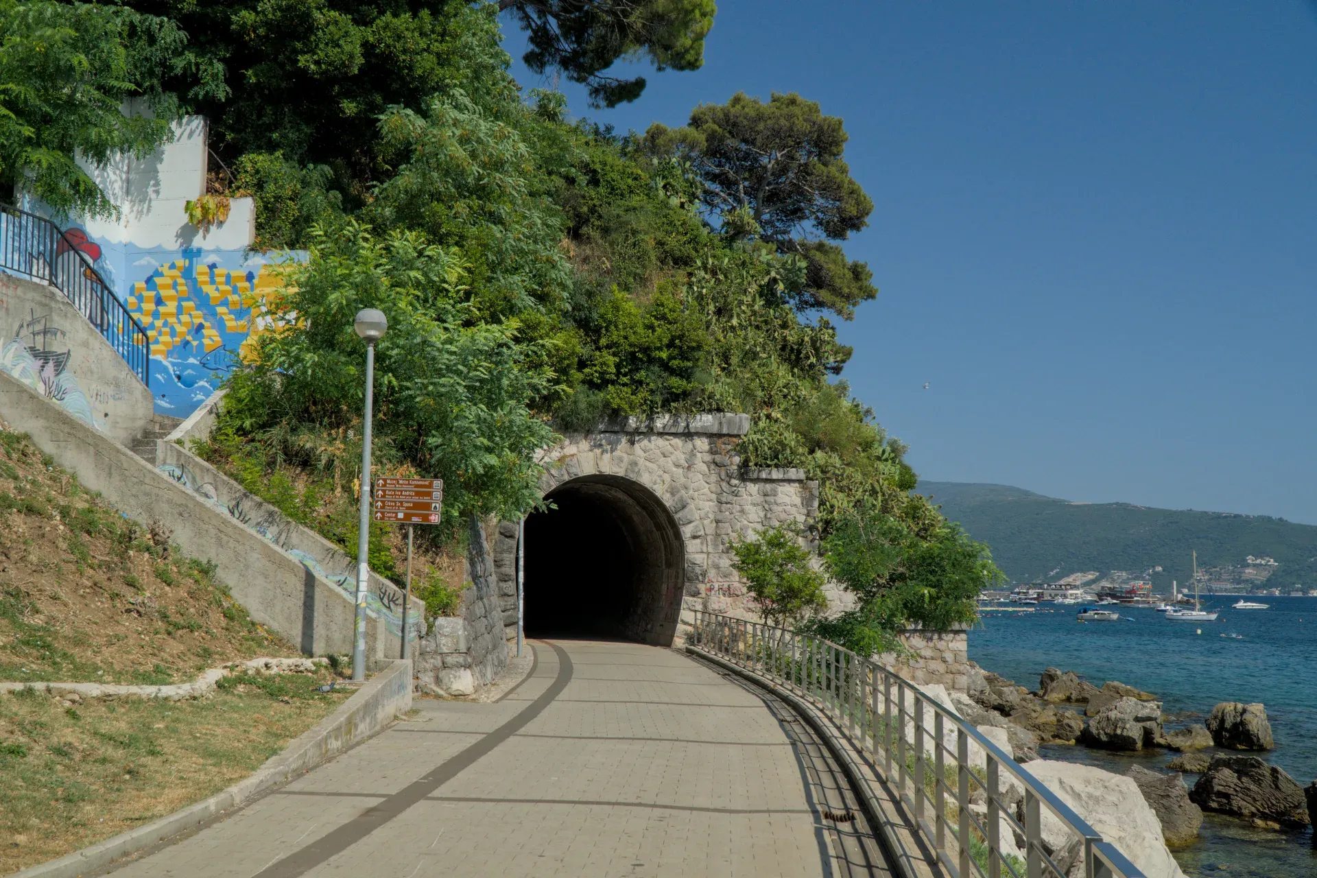 A stone tunnel with a lit entrance leads along the coast of Herceg Novi, Montenegro, overlooking the sea and mountains.