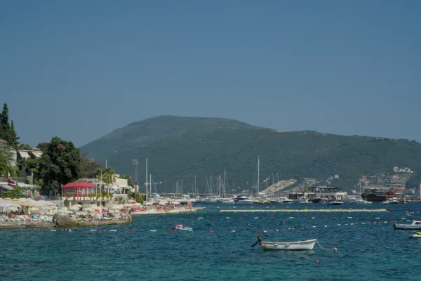Barcos en el puerto de Herceg Novi, Montenegro, con un animado paseo marítimo y colinas verdes al fondo.