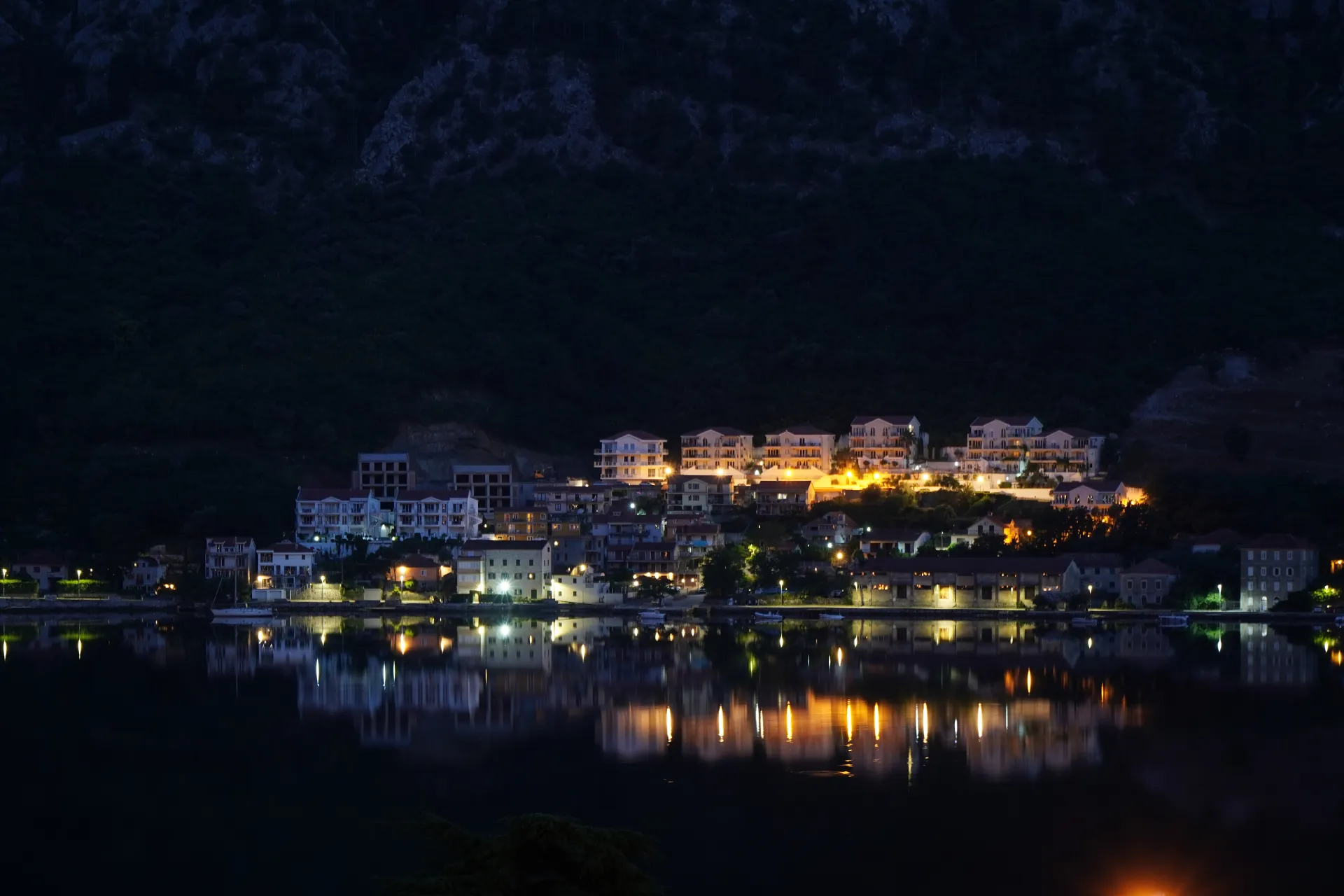 View of Kotor, Montenegro at night, showing illuminated buildings and their reflections in the bay.