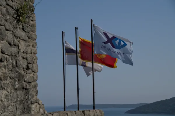 Tres banderas ondean al viento sobre el mar en Herceg Novi, junto a un antiguo muro de piedra.