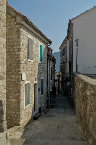 Un estrecho callejón adoquinado en Herceg Novi con casas de piedra antiguas y contraventanas verdes, a la luz del sol.