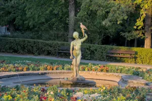 Bronzestatue eines Mädchens mit Füllhorn und Taube im Donaupark Novi Sad, Serbien, mit Blumen und Bäumen im Hintergrund.