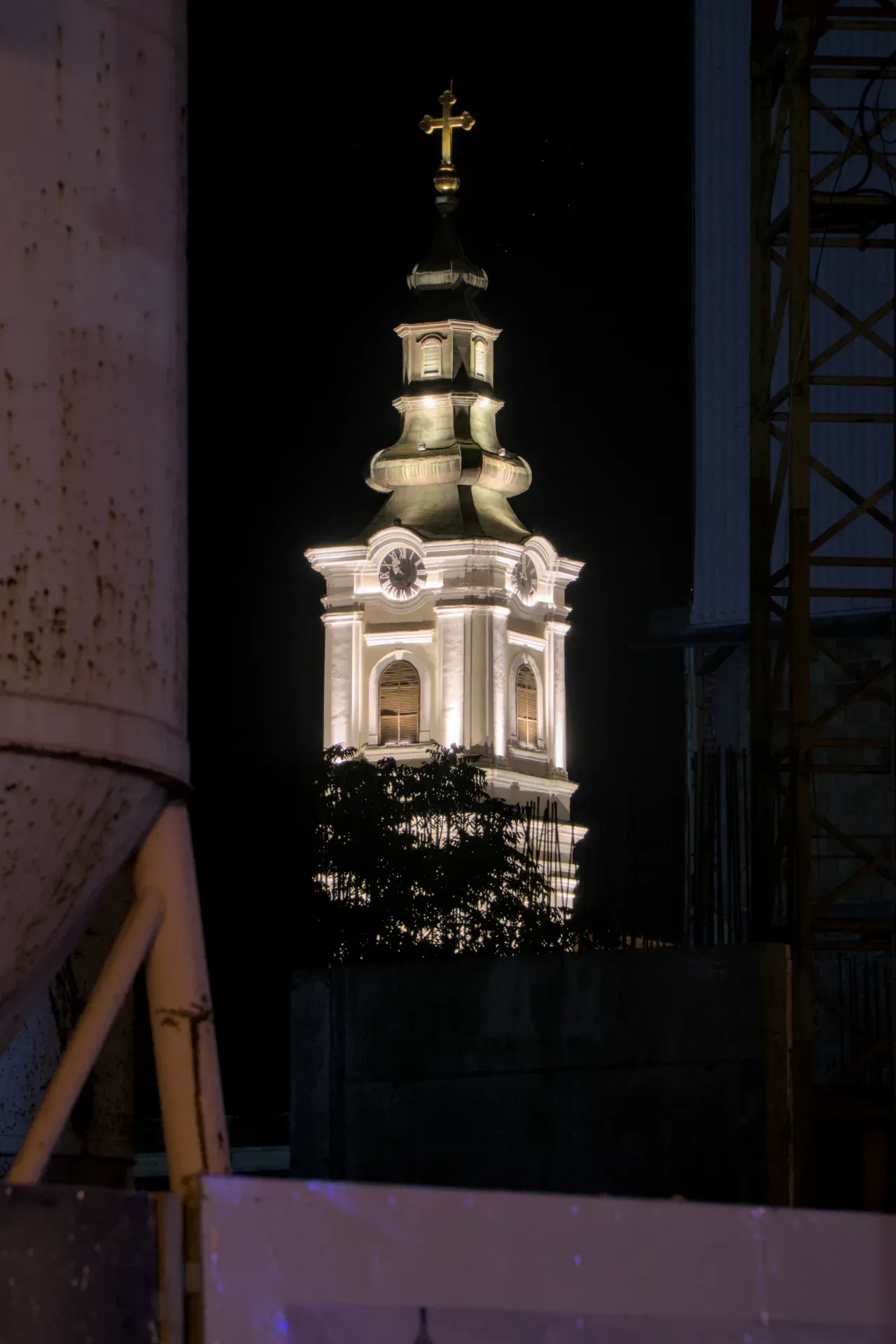 The illuminated bell tower of the Assumption of Mary Church in Novi Sad at night, framed by a dark sky and modern construction.