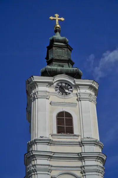 Zvonik Hrama Uspenja Presvete Bogorodice u Novom Sadu sa zlatnim krstom i satom, pod plavim nebom.