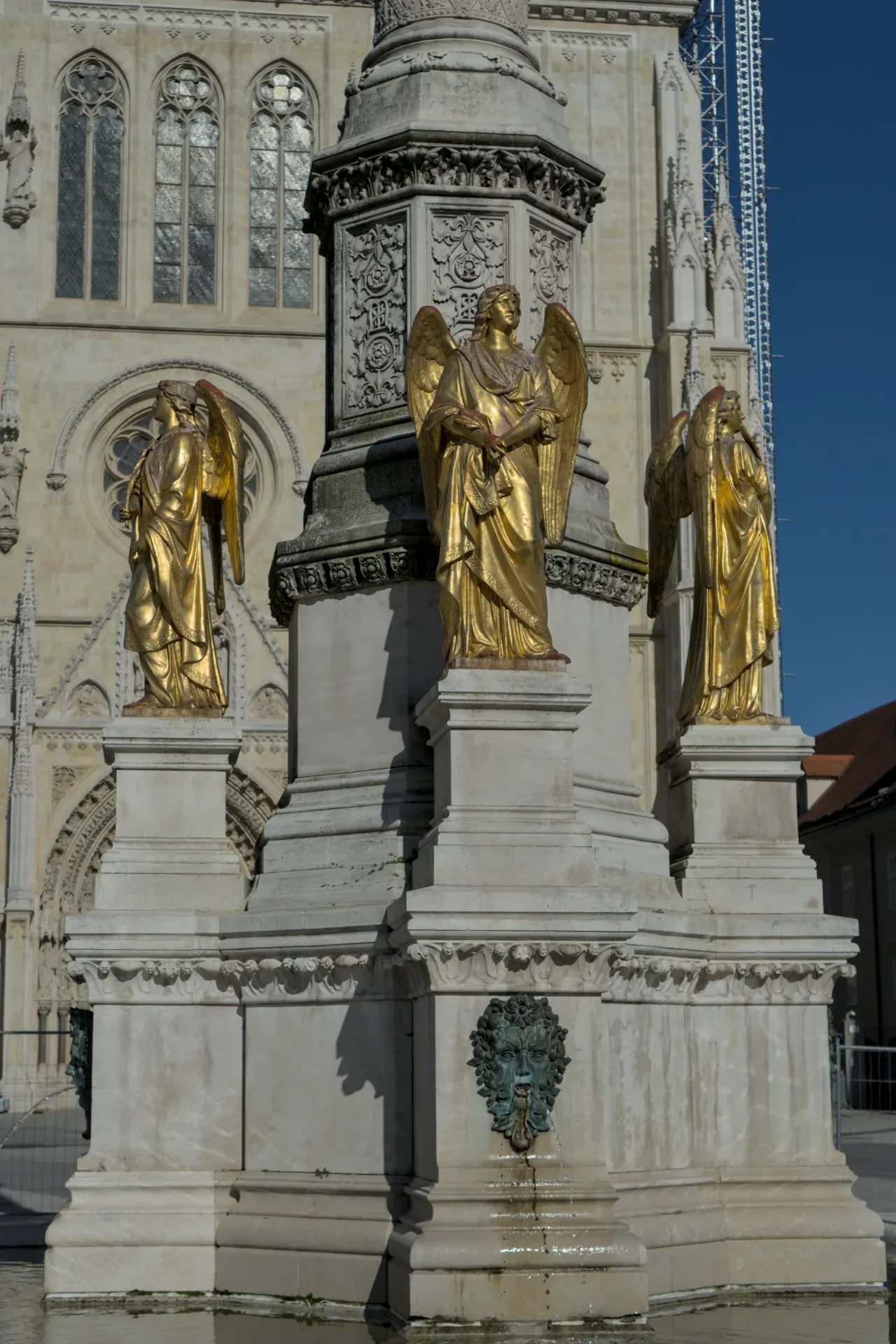 Three golden angel statues stand on a stone pillar in front of the monumental Zagreb Cathedral.