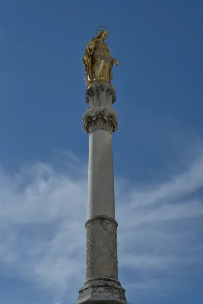 Statua dorata della Vergine Maria su una colonna finemente decorata, contro un cielo blu con nuvole sottili.