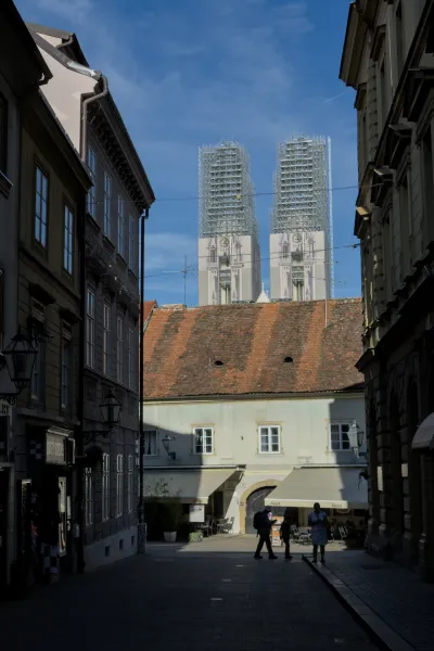 Architectural photograph of Zagreb Cathedral standing in scaffolding against a blue sky, with people walking through an alley.