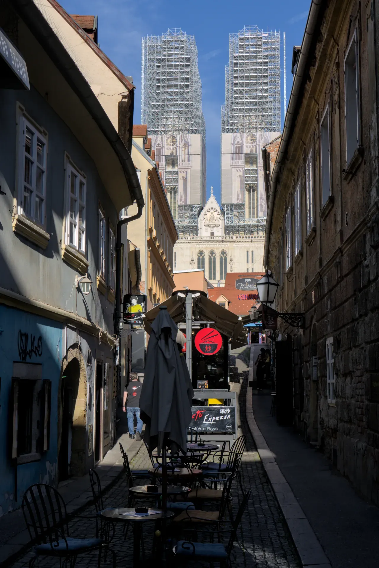 The impressive Zagreb Cathedral, partially obscured by construction scaffolding, rises above a charming narrow street with cafe tables and facades in the foreground.