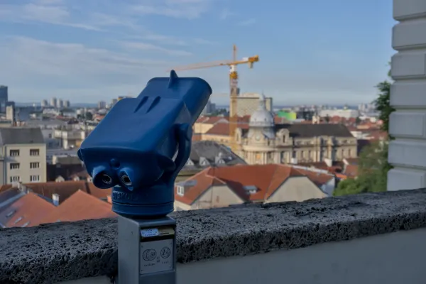 Un binocolo da osservazione blu si staglia davanti allo scenografico skyline di Zagabria, con tetti rossi e una gru sullo sfondo.