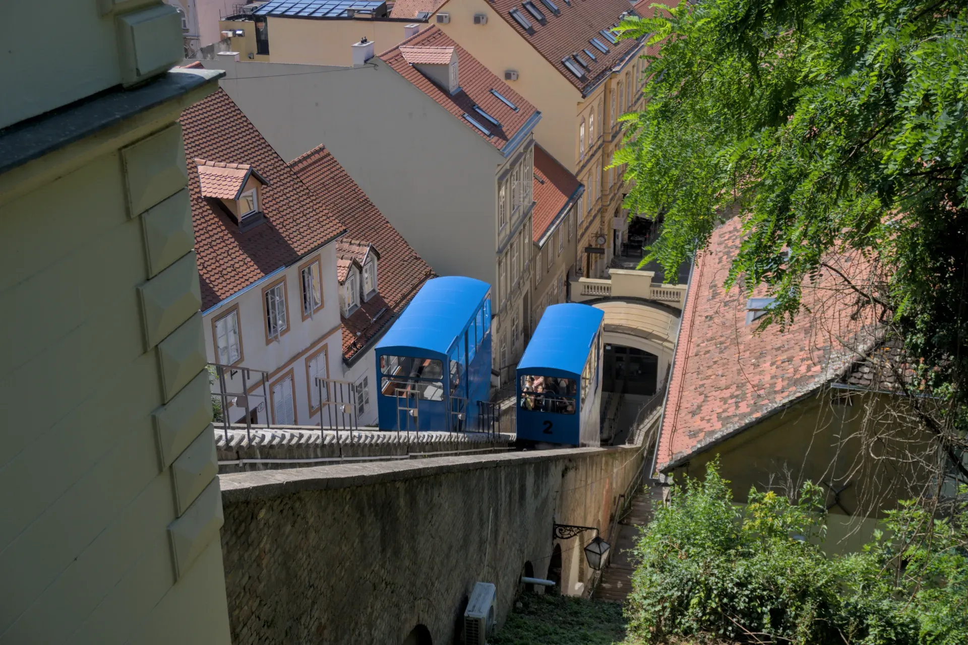 Two blue Zagreb funicular cars ascend a slope on a sunny day, surrounded by historic buildings and trees.