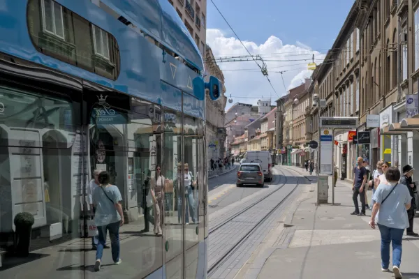 Un tramway bleu se déplace dans la rue animée de Zagreb, reflétant les bâtiments et les passants, avec une architecture historique en arrière-plan.
