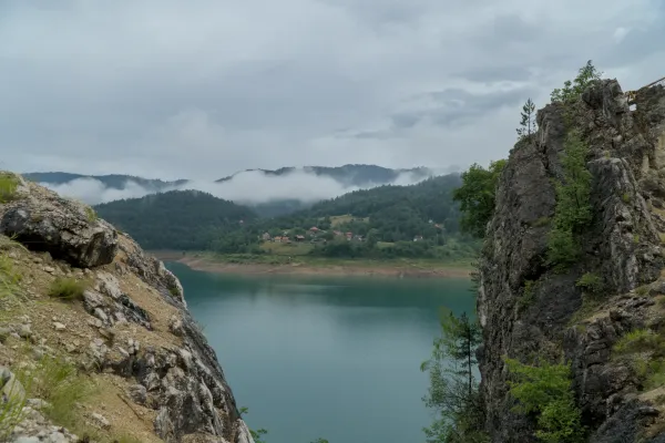 Панорама озера Заовине между скалистыми утесами, с лесистыми холмами и туманом на заднем плане.