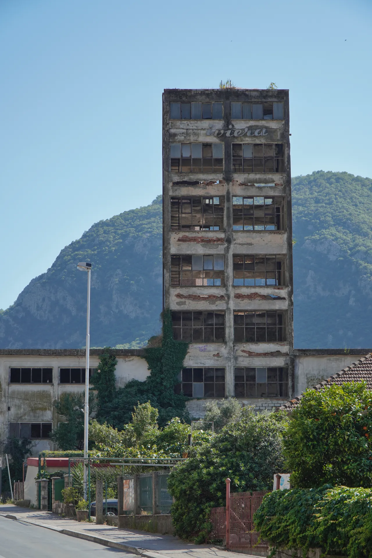 A tall, weathered, multi-story concrete building with broken windows and the word 'Riviera' painted on top, surrounded by greenery and mountains in Kotor, Montenegro.