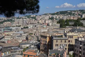 A wide view of Genoa, Italy, showing many buildings with terracotta roofs, some taller modern structures, and green hills in the background under a blue sky with white clouds.