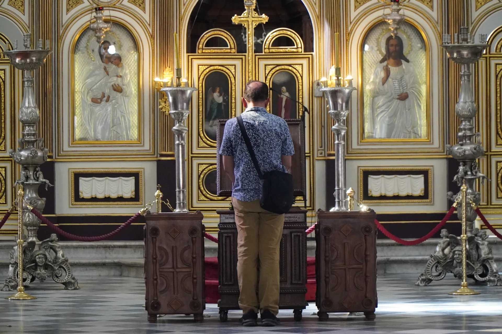 A man with his back to the viewer stands in an Orthodox church in Kotor, Montenegro, facing the ornate iconostasis adorned with religious icons and golden decorations.