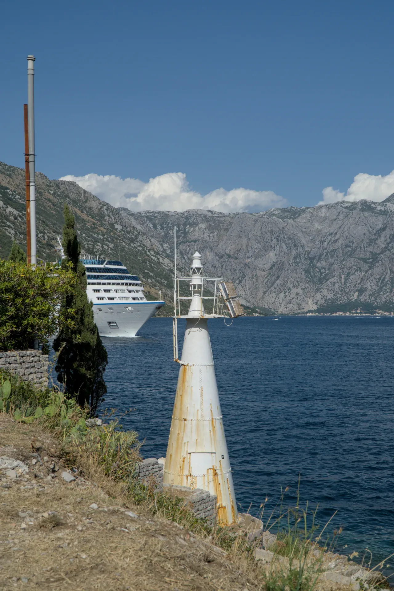 A weathered white lighthouse stands on the shore of Kotor Bay, with a large cruise ship sailing on the water.