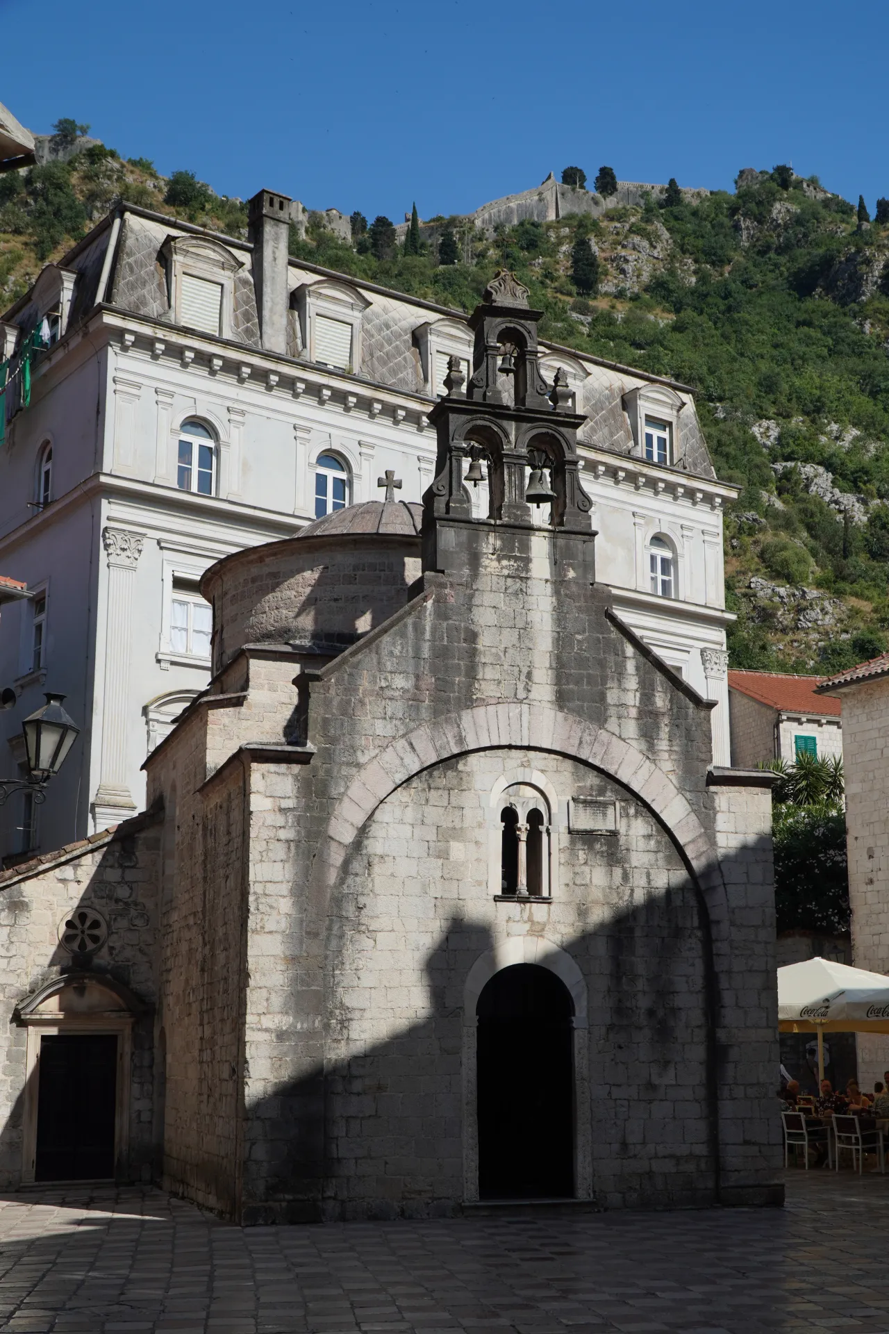 A view of St. Luke's Church in Kotor, Montenegro, featuring its stone architecture and bell tower, with the dramatic fortress walls of San Giovanni rising behind it.