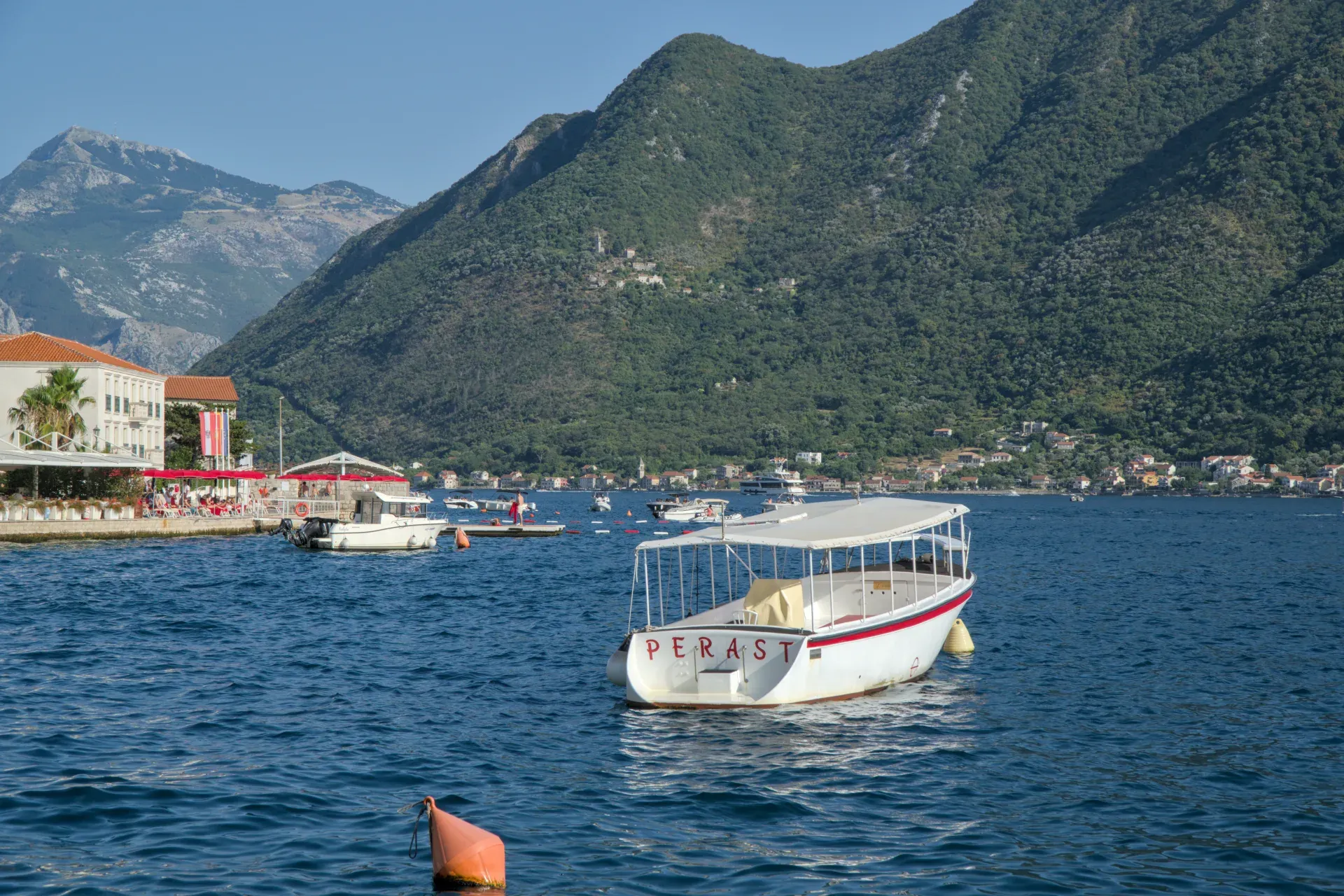 A small excursion boat named "Perast" anchors calmly in the Bay of Kotor, Montenegro, off the coast.