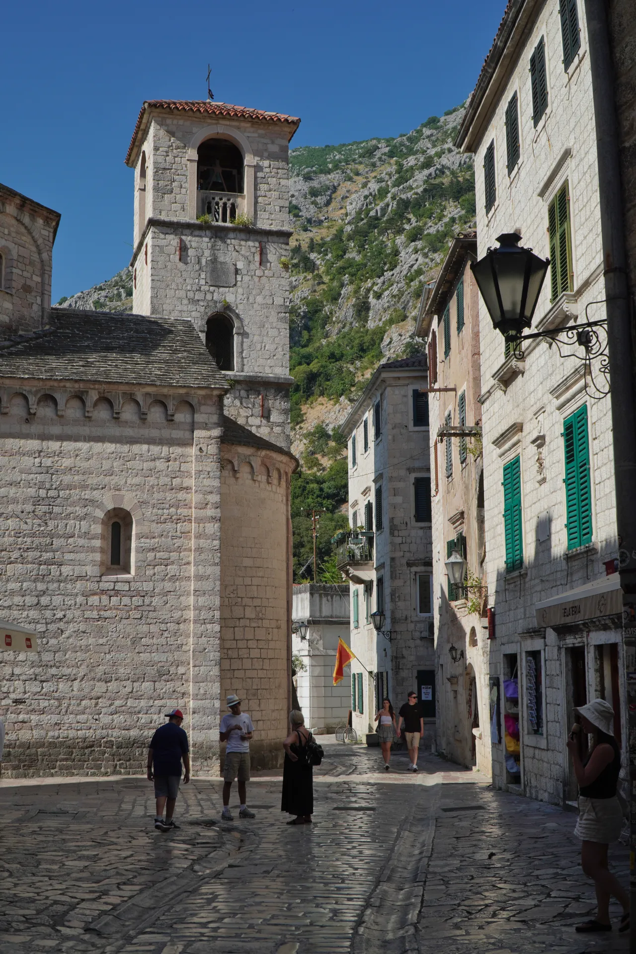 A street in Kotor Old Town with a stone bell tower, historic buildings, cobblestone pavement, and tourists.