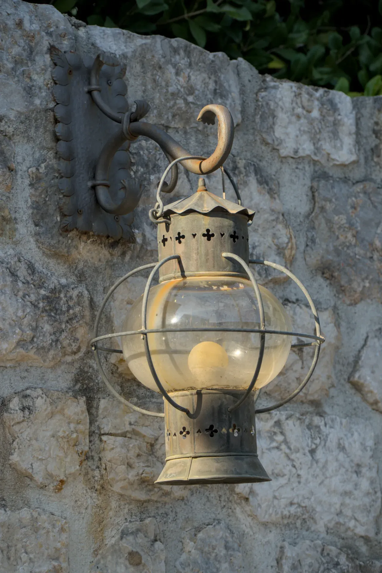 An antique, round lantern made of metal and glass hangs on a stone wall in Perast.