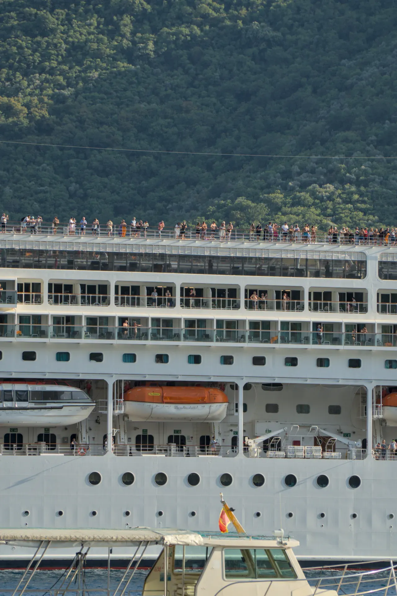 Passengers on the upper deck of a cruise ship wave goodbye to Kotor with a green mountain background.