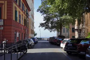 A sun-drenched street in Genoa, Italy, lined with colorful buildings with green shutters, parked cars and motorcycles, and a view of the blue Mediterranean Sea in the distance.