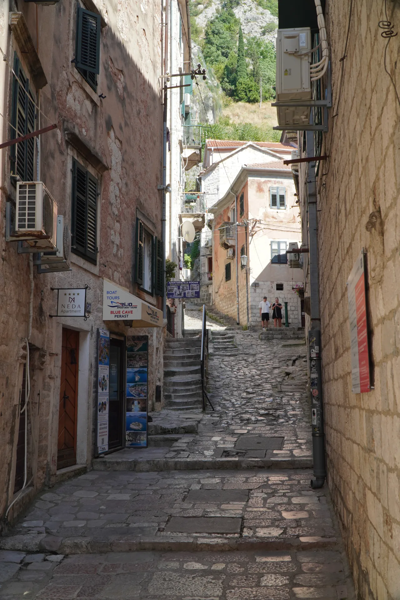 A steep, ancient cobblestone street in Kotor, Montenegro, lined with old stone buildings and leading upwards towards a green mountainside.