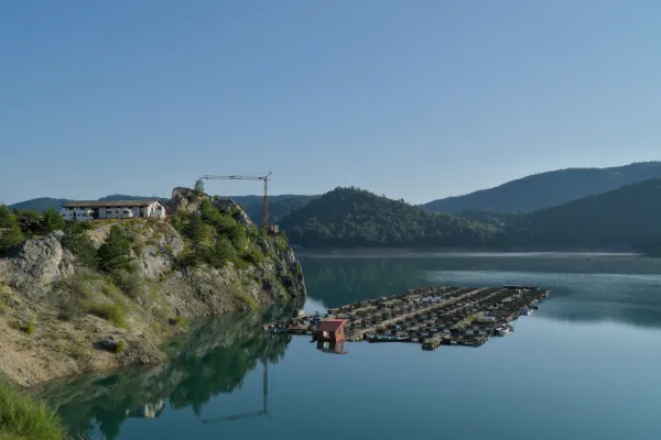 Amanecer en el Lago Zaovine, Serbia. Aguas tranquilas reflejan montañas y cielo azul con estructuras flotantes.