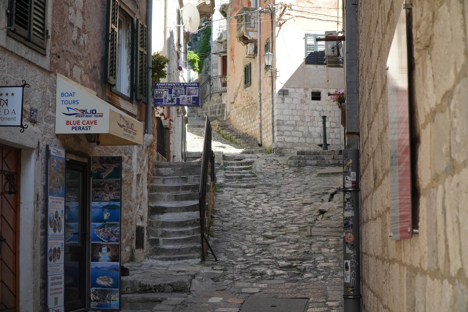 A narrow, stone-paved alleyway in Kotor's Old Town, Montenegro, with ancient buildings on either side, a set of stone stairs leading upwards, and signs advertising boat tours.